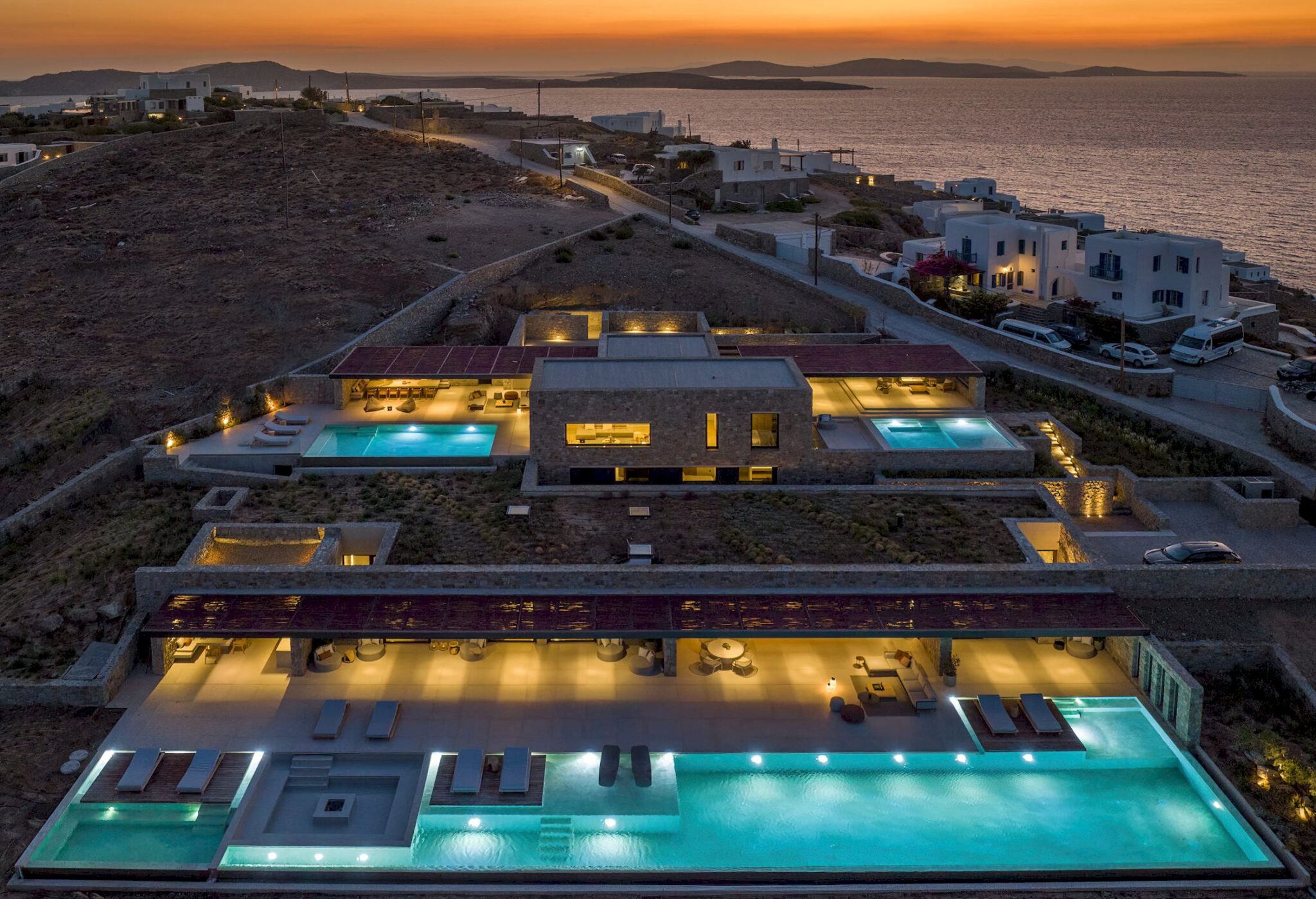Aerial view of illuminated villas and a long swimming pool overlooking the Mykonos coast at sunset