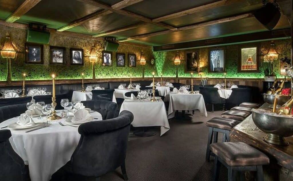 Restaurant interior at La Mangeoire Courchevel with red upholstered chairs, white tablecloths and an illuminated bar.